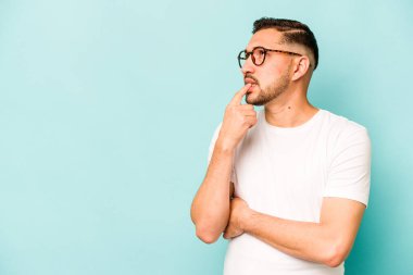 Genç İspanyol adam mavi arka planda izole edilmiş şüpheli ve şüpheci bir ifadeyle yana bakıyor..