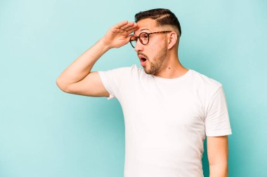 Genç İspanyol adam mavi arka planda izole edilmiş, uzaklara bakıyor ve elini alnına koyuyor..