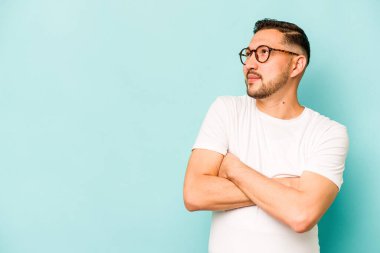 Mavi arka planda izole edilmiş genç İspanyol adam kollarını kavuşturarak gülümsüyor..