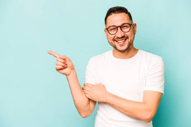 Mavi arka planda izole edilmiş genç İspanyol adam işaret parmağıyla işaret ederek gülümsüyor..