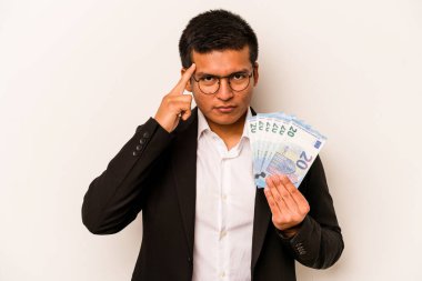 Latin kökenli genç bir iş adamı beyaz arka planda izole edilmiş banknotlar tutuyor. Şakağı işaret ediyor, düşünüyor, bir göreve odaklanıyor..