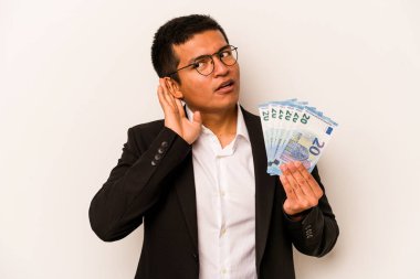 Latin kökenli genç bir iş adamı beyaz arka planda izole edilmiş banknotları tutuyor ve bir dedikoduyu dinlemeye çalışıyor..