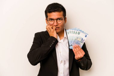 Latin kökenli genç bir iş adamı elinde banknotlar tutarken beyaz arka planda tırnak yiyor, gergin ve endişeli..