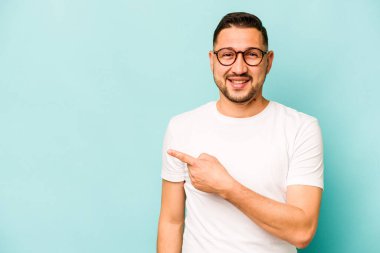 Mavi arka planda izole edilmiş genç İspanyol adam gülümsüyor ve boş alanda bir şeyler gösteriyor..