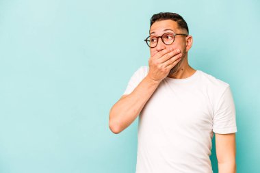 Genç İspanyol adam mavi arka planda izole edilmiş. Düşünceli bir şekilde bir fotokopi odasına bakıyor. Ağzını eliyle kapatıyor..