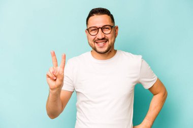 Mavi arka planda izole edilmiş genç İspanyol adam zafer işareti gösteriyor ve gülümsüyor..