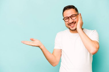Mavi arka planda izole edilmiş genç İspanyol bir adam avucunun içinde fotokopi alanı tutuyor, el üstünde tutuyor. Şaşırmış ve mutlu..