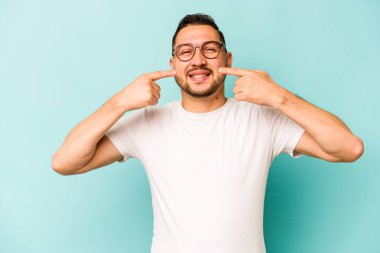 Mavi arka planda izole edilmiş genç İspanyol adam gülümsüyor, parmaklarını ağıza doğrultuyor..