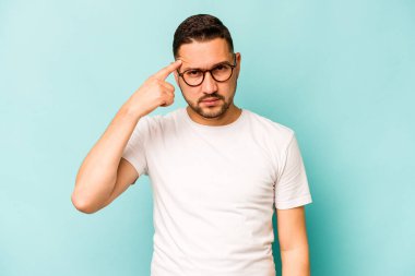 Genç İspanyol bir adam mavi arka planda izole edilmiş, parmağıyla tapınağı işaret ediyor, düşünüyor ve bir göreve odaklanıyor..