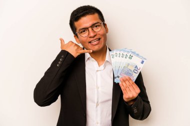 Latin kökenli genç bir iş adamı beyaz arka planda izole edilmiş banknotlar tutuyor ve parmakları olan bir cep telefonu hareketi gösteriyor..