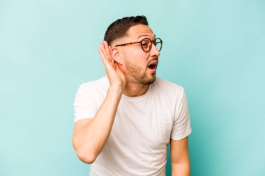 Genç İspanyol bir adam mavi arka planda izole edilmiş bir şekilde bir dedikoduyu dinlemeye çalışıyor..