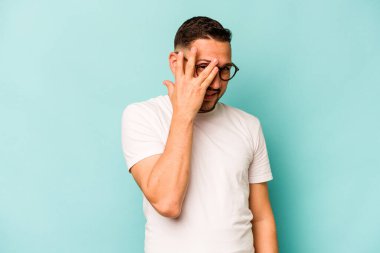 Mavi arka planda izole edilmiş genç İspanyol adam kameraya parmaklarıyla göz kırpıyor, yüzünü kapatmaktan utanıyor..