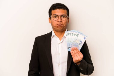 Latin kökenli genç bir iş adamı beyaz arka planda soyutlanmış banknotlar tutuyor omuz silkiyor ve gözleri açık bakıyor..