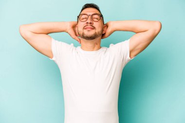 Genç İspanyol adam mavi arka planda izole edilmiş. Kendine güveniyor, elleri başının arkasında..