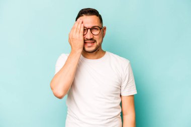 Genç İspanyol adam mavi arka planda izole edilmiş. Yüzünün yarısını avuç içiyle kaplayarak eğleniyor..