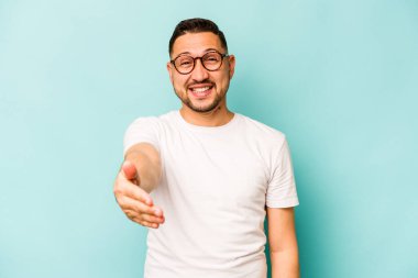 Mavi arka planda izole edilmiş genç İspanyol adam selamlama jesti olarak kameraya uzanıyor..