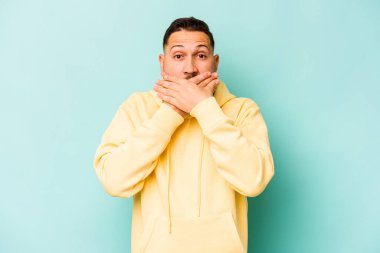 Mavi arka planda izole edilmiş genç İspanyol adam elleriyle ağzını kapatırken şok oldu..