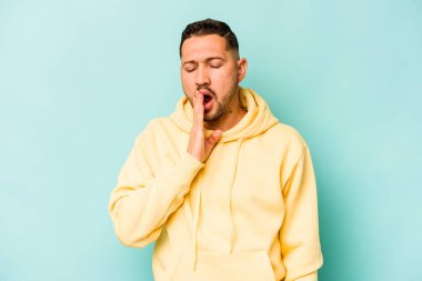 Mavi arka planda izole edilmiş genç İspanyol adam esniyor. Ağzı eliyle kapatan yorgun bir hareket sergiliyor..