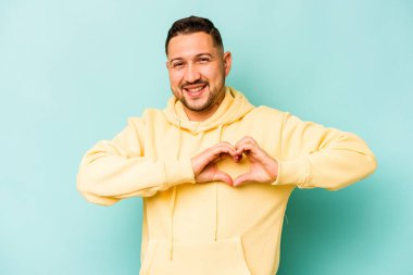 Mavi arka planda izole edilmiş genç İspanyol adam gülümsüyor ve elleriyle kalp şeklini gösteriyor..