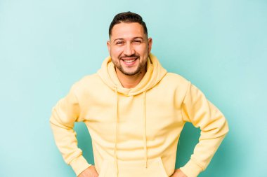 Latin kökenli genç adam mavi arka planda izole edilmiş. Kendine güveniyor. Kalçaları tutuyor..