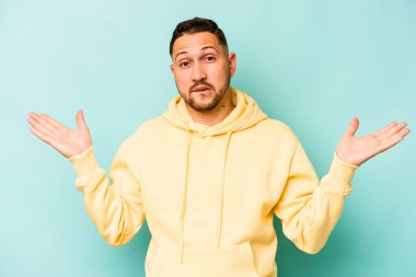 Genç İspanyol bir adam mavi arka planda izole edilmiş. Kafası karışmış ve omuz silkiyor. Kopya alanını tutmak için..