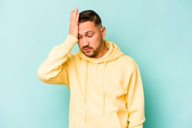 Genç İspanyol adam mavi arka planda izole edilmiş. Çok yorgun ve uykulu. Elini başının üstünde tutuyor..