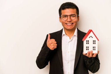 Latin kökenli genç bir adam beyaz arka planda izole edilmiş küçük bir ev tutuyor gülümsüyor ve başparmağını kaldırıyor.