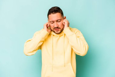 Mavi arka planda izole edilmiş genç İspanyol adam kulaklarını elleriyle kapatıyor..