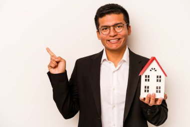 Beyaz arka planda izole edilmiş küçük bir evi tutan İspanyol genç adam gülümsüyor ve boş alanda bir şeyler gösteriyor..