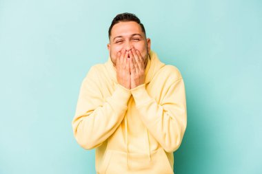 Genç İspanyol bir adam mavi arka planda izole edilmiş bir şekilde bir şeye gülüyor, elleriyle ağzını kapatıyor..