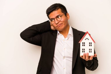 Latin kökenli genç bir adam beyaz arka planda izole edilmiş küçük bir ev tutuyor kafasının arkasına dokunuyor, düşünüyor ve bir seçim yapıyor..