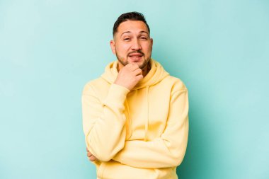 Genç İspanyol adam mavi arka planda izole edilmiş mutlu ve kendinden emin gülümsüyor, elleriyle çenesine dokunuyor..