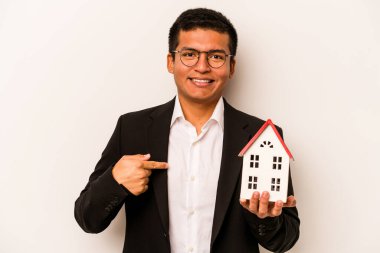 Latin kökenli genç bir adam, beyaz arka planda küçük bir ev tutuyor. Bir gömlek fotokopisini elle gösteriyor. Gururlu ve kendinden emin.