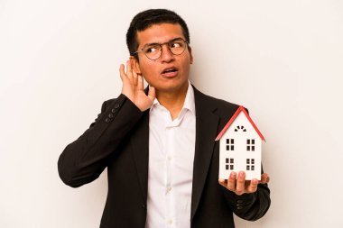 Latin kökenli genç bir adam beyaz arka planda küçük bir ev tutuyor ve bir dedikoduyu dinlemeye çalışıyor..