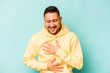 Mavi arka planda izole edilmiş genç İspanyol adam mutlu bir şekilde gülüyor ve ellerini karnının üstünde tutarak eğleniyor..
