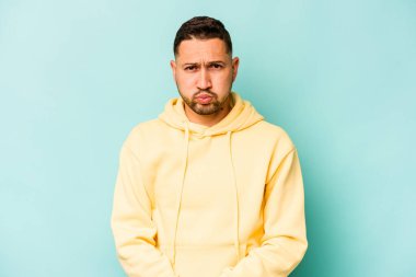 Mavi arka planda izole edilmiş genç İspanyol adam yanakları patlatıyor, yorgun bir ifadesi var. Yüz ifadesi kavramı.