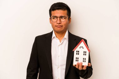 Latin kökenli genç bir adam beyaz arka planda küçük bir ev tutuyor. Kafası karışmış, kendinden emin değil..