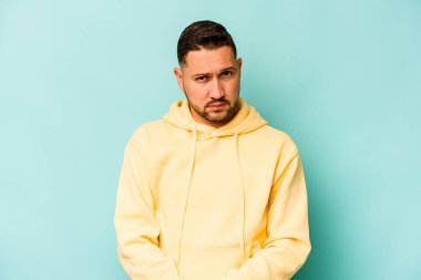 Latin asıllı genç adam mavi arka planda izole edilmiş. Mutsuz bir şekilde kameraya bakıyor. İğneleyici bir ifadeyle..