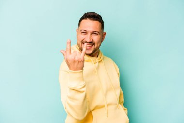 Mavi arka planda izole edilmiş genç İspanyol adam parmaklarıyla kaya hareketi gösteriyor.