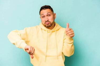 Latin kökenli genç adam mavi arka planda izole edilmiş, baş parmağını kaldırıyor, zor bir seçim konsepti.