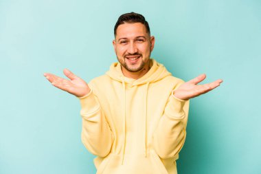 Mavi arka planda izole edilmiş genç İspanyol adam kollarıyla ölçeği yapıyor, mutlu ve kendinden emin hissediyor..