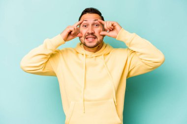 Genç İspanyol bir adam mavi arka planda izole edilmiş. Başarı fırsatı bulmak için gözlerini açık tutuyor..