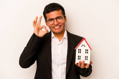 Latin kökenli genç bir adam beyaz arka planda izole edilmiş küçük bir ev tutuyor. Neşeli ve kendinden emin..