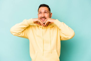 Mavi arka planda izole edilmiş genç İspanyol adam ellerini çenesinin altında tutuyor ve mutlu bir şekilde yan tarafa bakıyor..