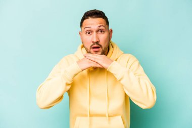 Latin kökenli genç adam mavi arka planda izole edilmiş şans için dua ediyor, şaşırmış ve ağzı açık bir şekilde önüne bakıyor..