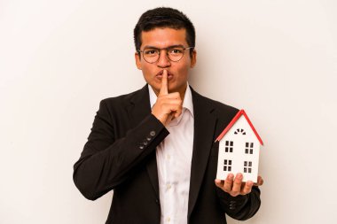 Latin kökenli genç bir adam beyaz arka planda izole edilmiş küçük bir ev tutuyor ya sır saklıyor ya da sessizlik istiyor..