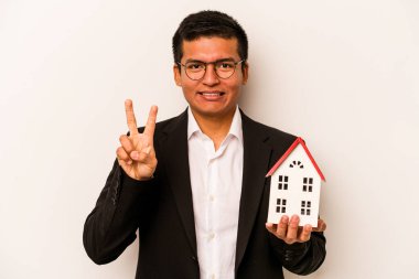 Beyaz arka planda küçük bir ev tutan İspanyol genç adam iki numarayı parmaklarıyla gösteriyor..