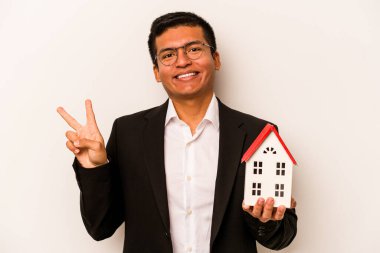 Genç İspanyol iş adamı beyaz arka planda izole edilmiş küçük bir ev tutuyor neşeli ve kaygısız parmaklarıyla barış sembolü gösteriyor..