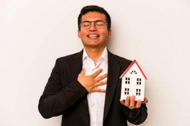 Beyaz arka planda izole edilmiş küçük bir evi tutan İspanyol genç adam kahkahalarla gülüyor ve elini göğsünde tutuyor..