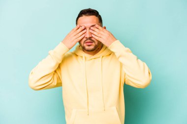 Mavi arka planda izole edilmiş genç İspanyol adam elleriyle gözlerini kapatmaktan korkuyor..